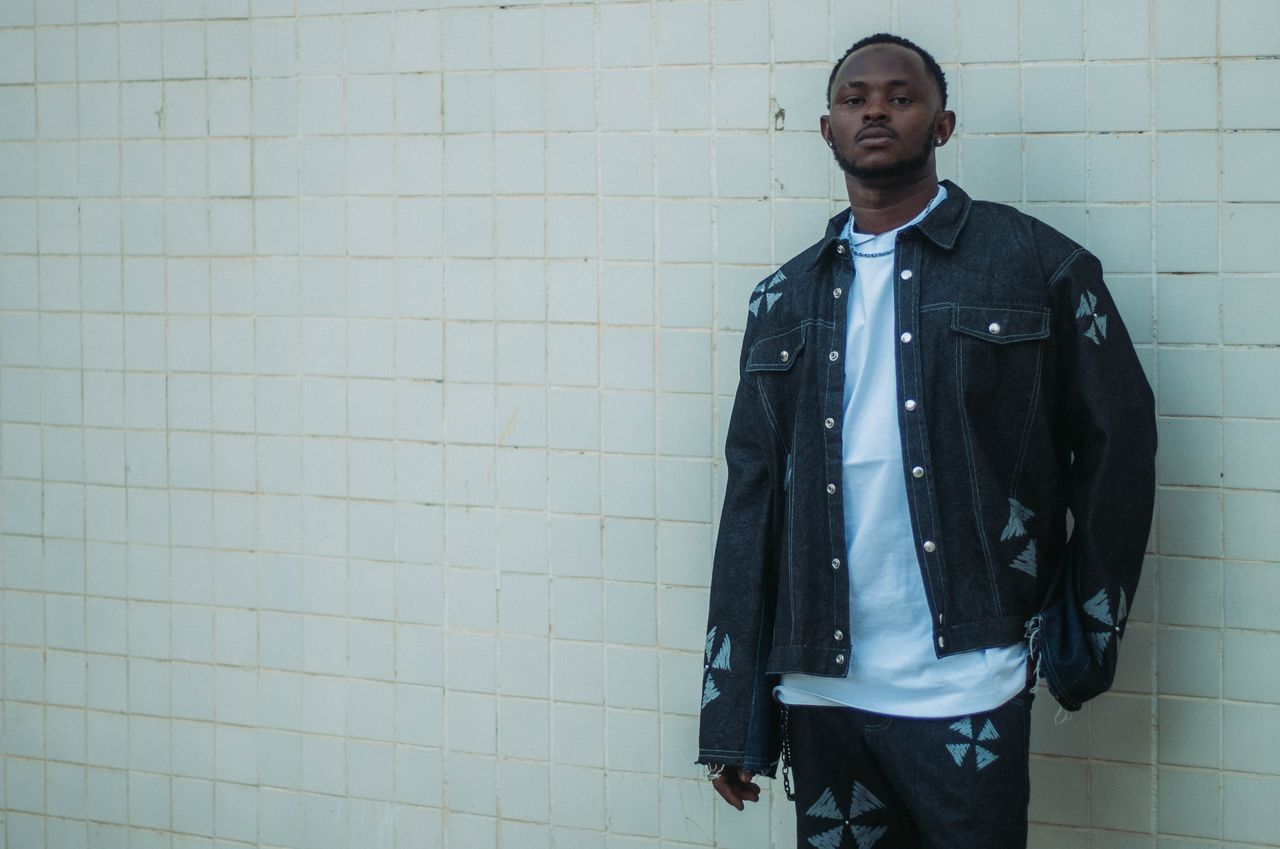 Fashionable man in a dark denim jacket and jeans poses against a tiled wall.